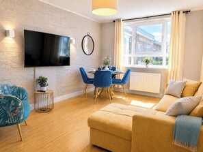 Ein modernes Wohnzimmer mit Flachbildfernseher, einem Essbereich mit blauen Stühlen, einer Couch und einem Fenster mit Vorhängen.