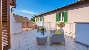 A balcony with a tiled floor, two white chairs with yellow cushions, a round table, and a building with green shutters in the background.