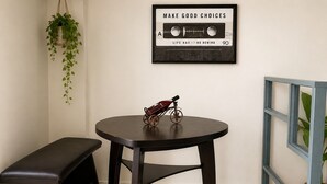 Ein Zimmer mit einem runden Tisch, einer Bank, einem gerahmten Motivationszitat, einer hängenden Pflanze und einem Fenster mit blauem Rahmen.