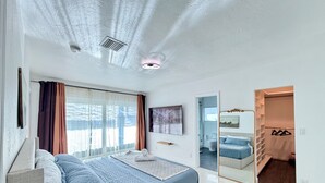 Quarto moderno com cama, mesa de cabeceira, janela com cortinas, banheiro visível através de uma porta aberta e armário com roupas penduradas.