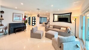 Ampla sala de estar com um piano, conjunto de sofás, mesa de jantar e uma grande janela com persianas.