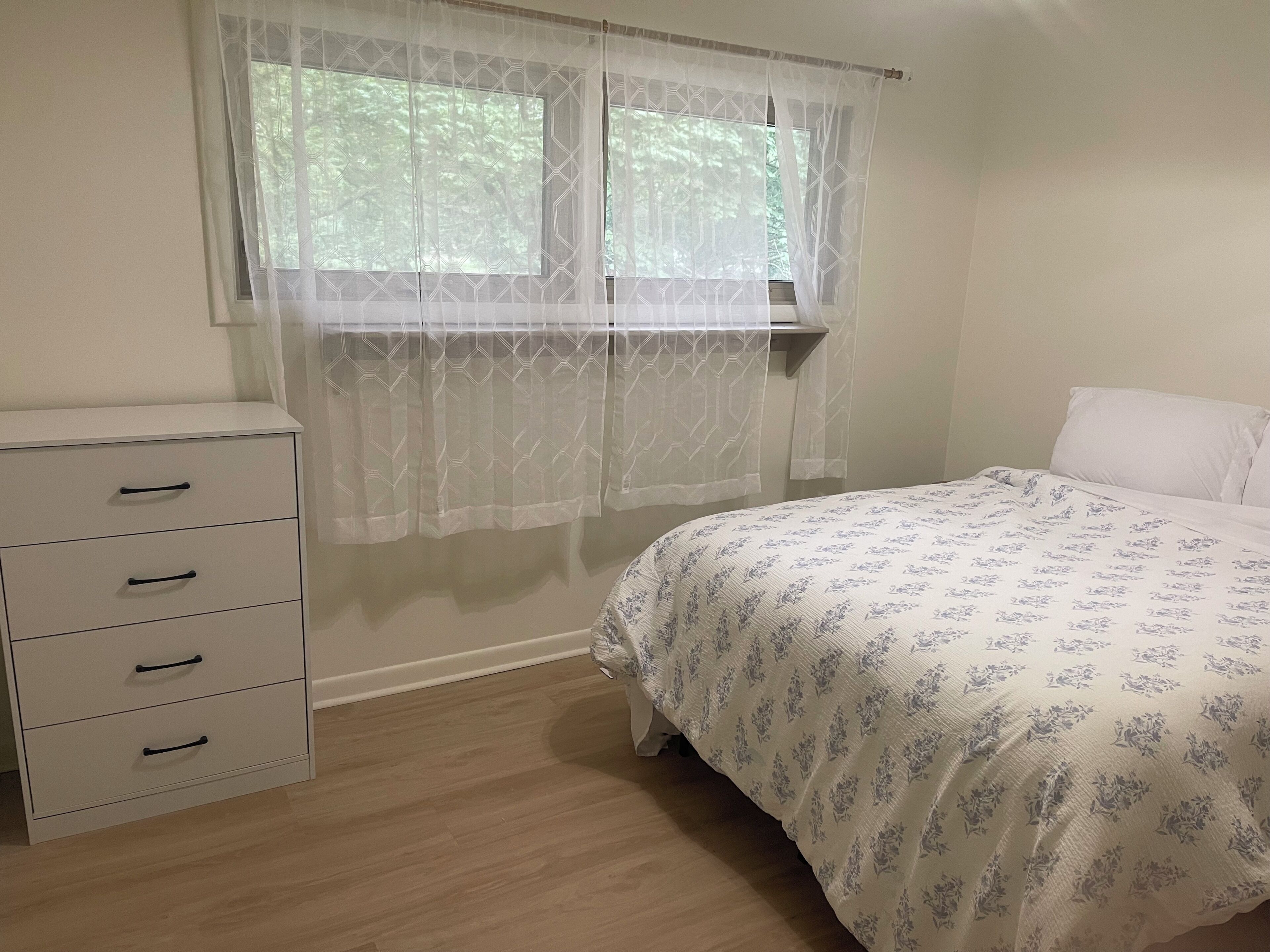 Ein Schlafzimmer mit einem Bett, einer Kommode und einem Fenster mit durchsichtigen Vorhängen.
