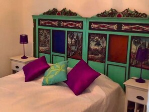 Ein Bett mit weißer Bettdecke, zwei violette Kissen und ein türkisblaues Kissen mit gelbem Muster.