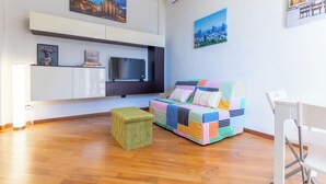 Een moderne woonkamer met een flatscreen televisie, een kleurrijke bank, een klein poefje en kunstwerken aan de muren.