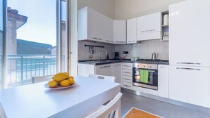 Een moderne keuken met witte kastjes, een eettafel met een schaal fruit, en uitzicht door een groot raam.