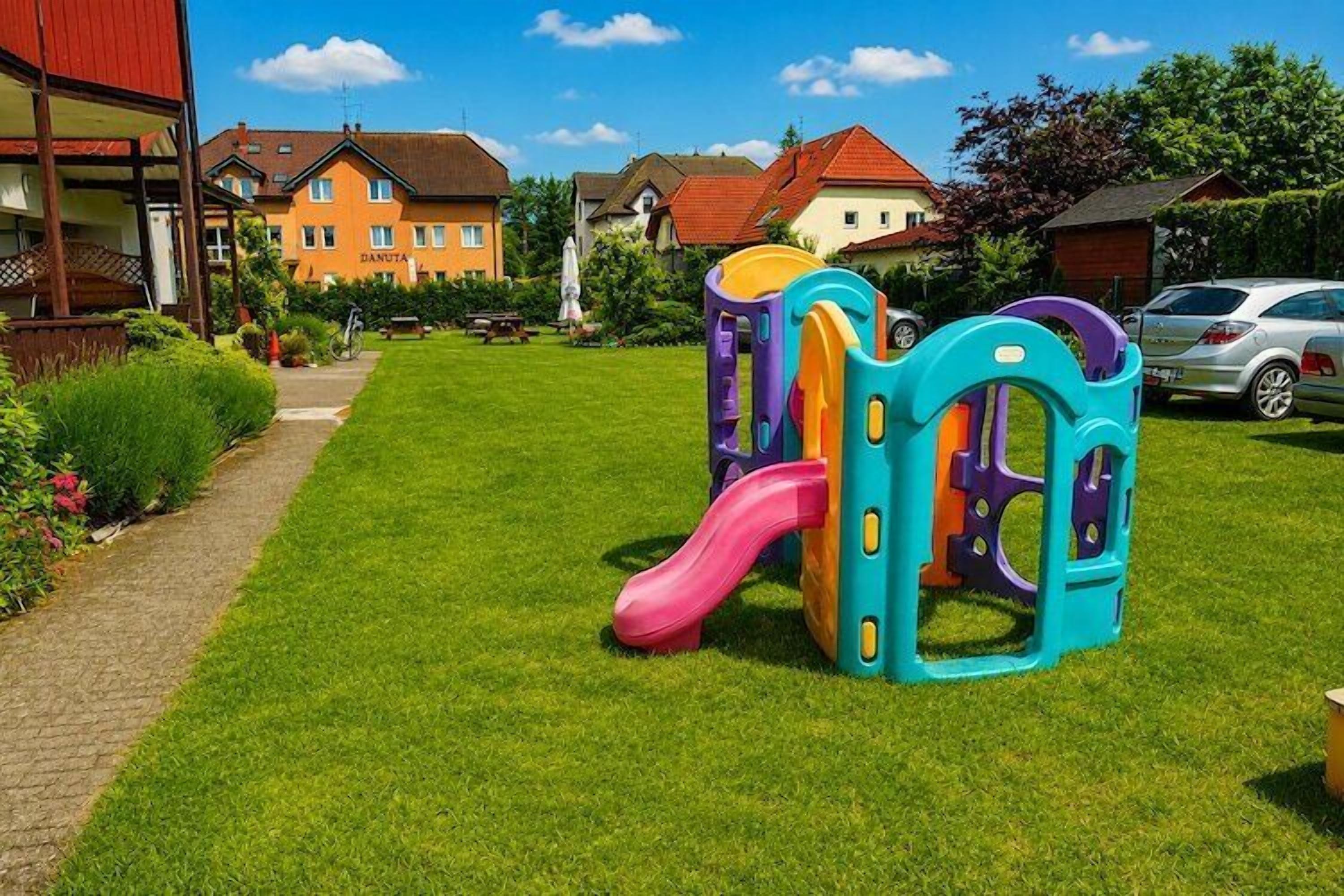 Un terrain de jeux avec un toboggan et des structures d’escalade, situé dans un espace verdoyant, avec des maisons en arrière-plan.