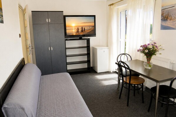 A compact room with a bed, a television mounted on a cabinet, a dining table with chairs, and a window with curtains.