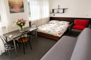 A room with a bed, a desk, a chair, a sofa, a window with curtains, a wall-mounted shelf with decorative items, and a painting on the wall.