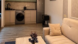 Een moderne keuken met groene kastjes, een wasmachine en een koelkast. Er staat een beige bank en een kleine tafel met een vaas.