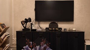 Een moderne woonkamer met een flatscreen televisie aan de muur, een donkere houten entertainmentkast met elektronische apparaten, een witte vaas met paarse bloemen op een houten tafel, en een boekenplank vol boeken.