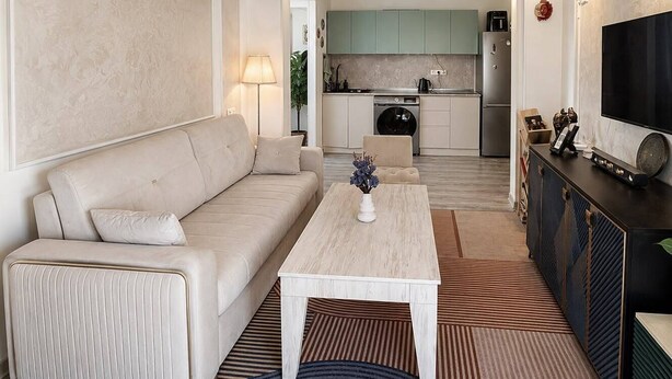 Een moderne woonkamer met een bank, een salontafel en een televisie. Achtergrond is een keuken met kasten en apparatuur.