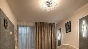 Een slaapkamer met een bed, nachtkastje, wandklok, raam met gordijnen en een plafondlamp.