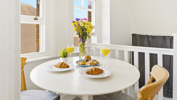 Une table ronde blanche, recouverte d’assiettes remplies de nourriture, un verre de jus d’orange et un vase de fleurs, le tout sur un balcon.