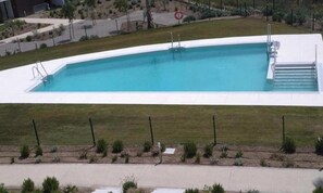Une piscine rectangulaire avec une plage blanche, entourée d’une clôture et d’une zone herbeuse.