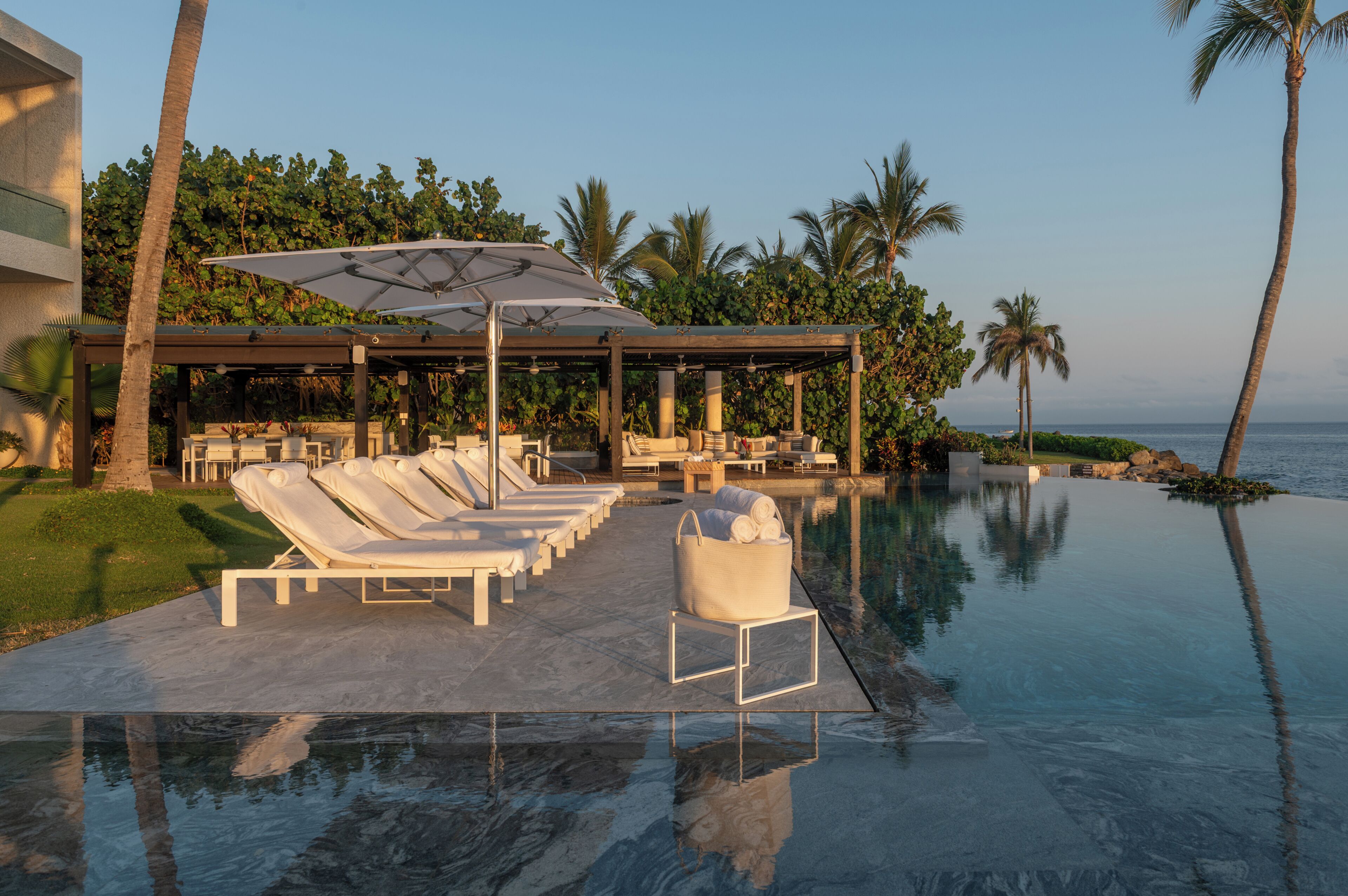 An infinity pool with lounge chairs and umbrellas, surrounded by palm trees and a clear view of the ocean.