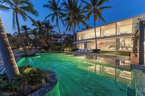 A modern resort with a swimming pool, palm trees, and a two-story building with glass balconies. - Breeze 5-18 | Oceanfront Villa | Infinity Pool (Cruz de Huanacaxtle)