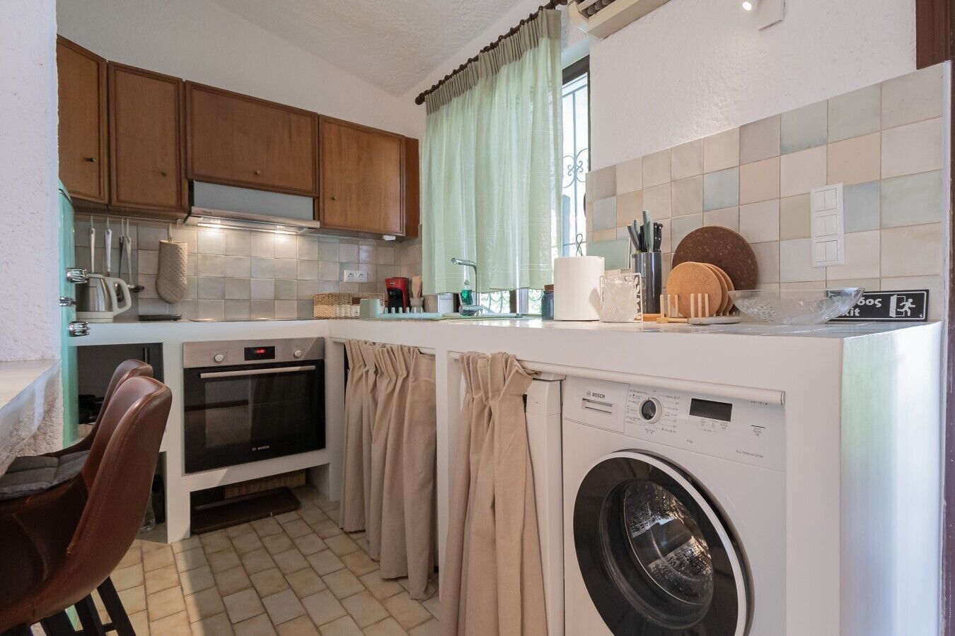 A compact kitchen with wooden cabinets, a washing machine, a coffee maker, and a dining area with a chair.
