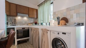 Een compacte keuken met houten kastjes, een wasmachine, een koffiezetapparaat en een eethoek met een stoel.