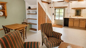 Una habitación con una mesa de comedor de madera, dos sillones a rayas, una escalera, una cocina con armarios de madera y una puerta blanca.