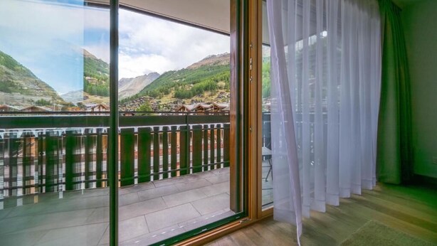 Ein Zimmer mit einer großen Schiebetür, die auf einen Balkon mit Blick auf grüne Hügel und ein Gewässer führt.