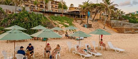 A beach with white lounge chairs and green umbrellas, palm trees, and buildings in the background.