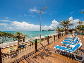 Terraza de madera con sillones de descanso frente a la playa, una valla y palmeras.