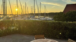 Um pátio com uma mesa de madeira redonda e quatro cadeiras, com vista para uma marina com vários veleiros e mastros.