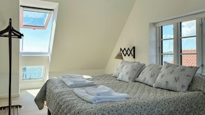 Quarto com teto inclinado, cama com roupa de cama floral, duas janelas e uma luminária fixada na parede.