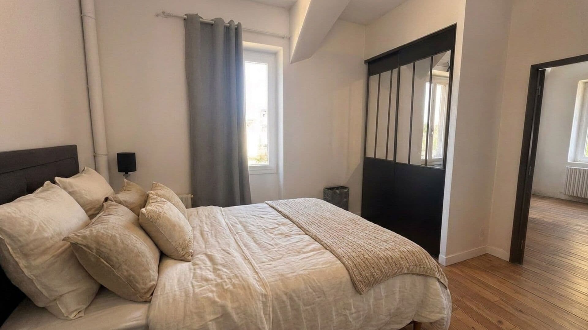 Ein Schlafzimmer mit einem Bett, Kissen, einer Nachttischlampe, einem Fenster mit Vorhängen und einer Tür mit schwarzem Rahmen.