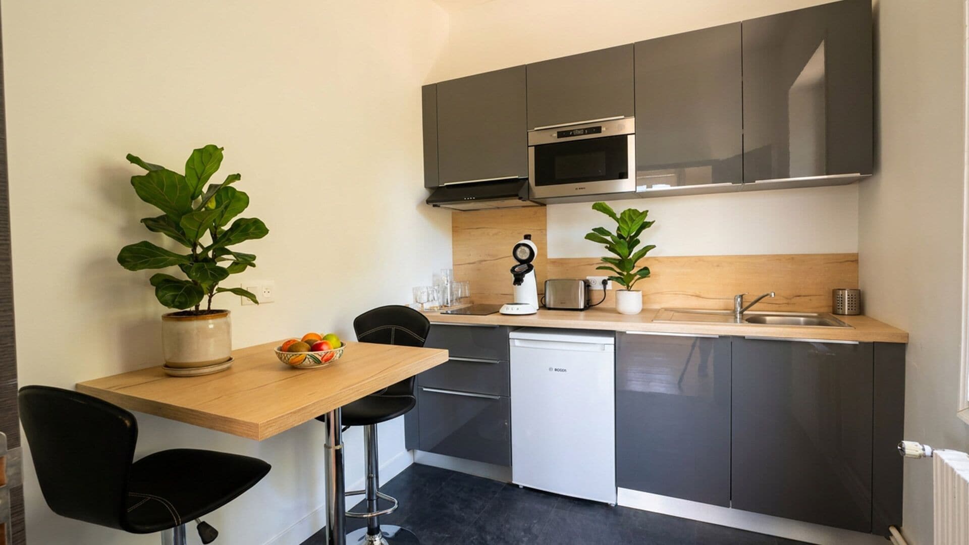 A compact kitchenette with a microwave, toaster, and sink. There is a small dining area with a table and chairs, and two potted plants.