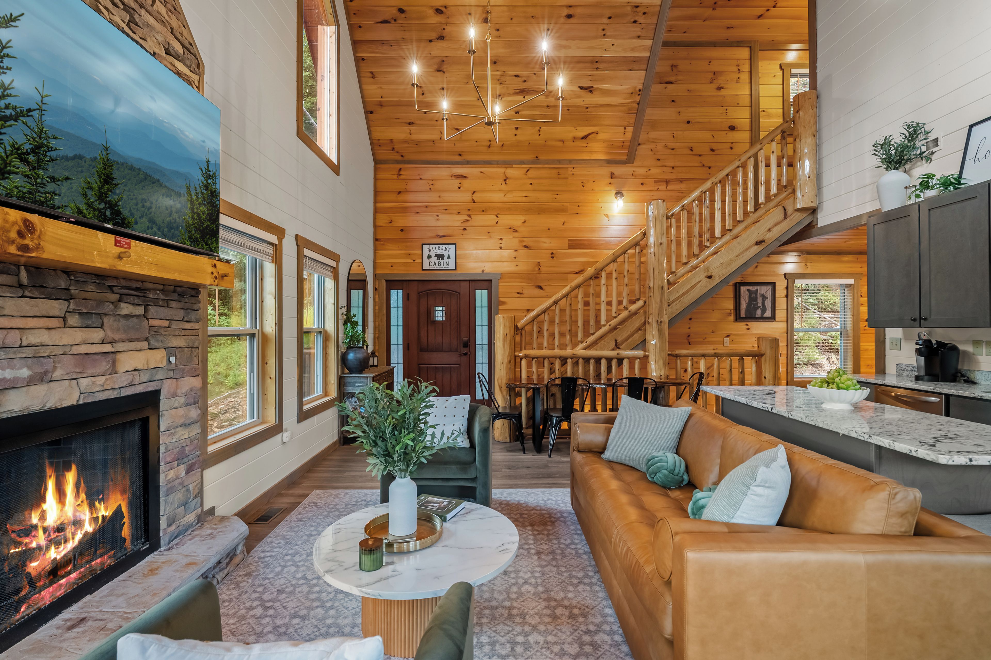 A cabin interior with a fireplace, wooden ceiling, a staircase, a living area with a sofa, and a kitchen area.