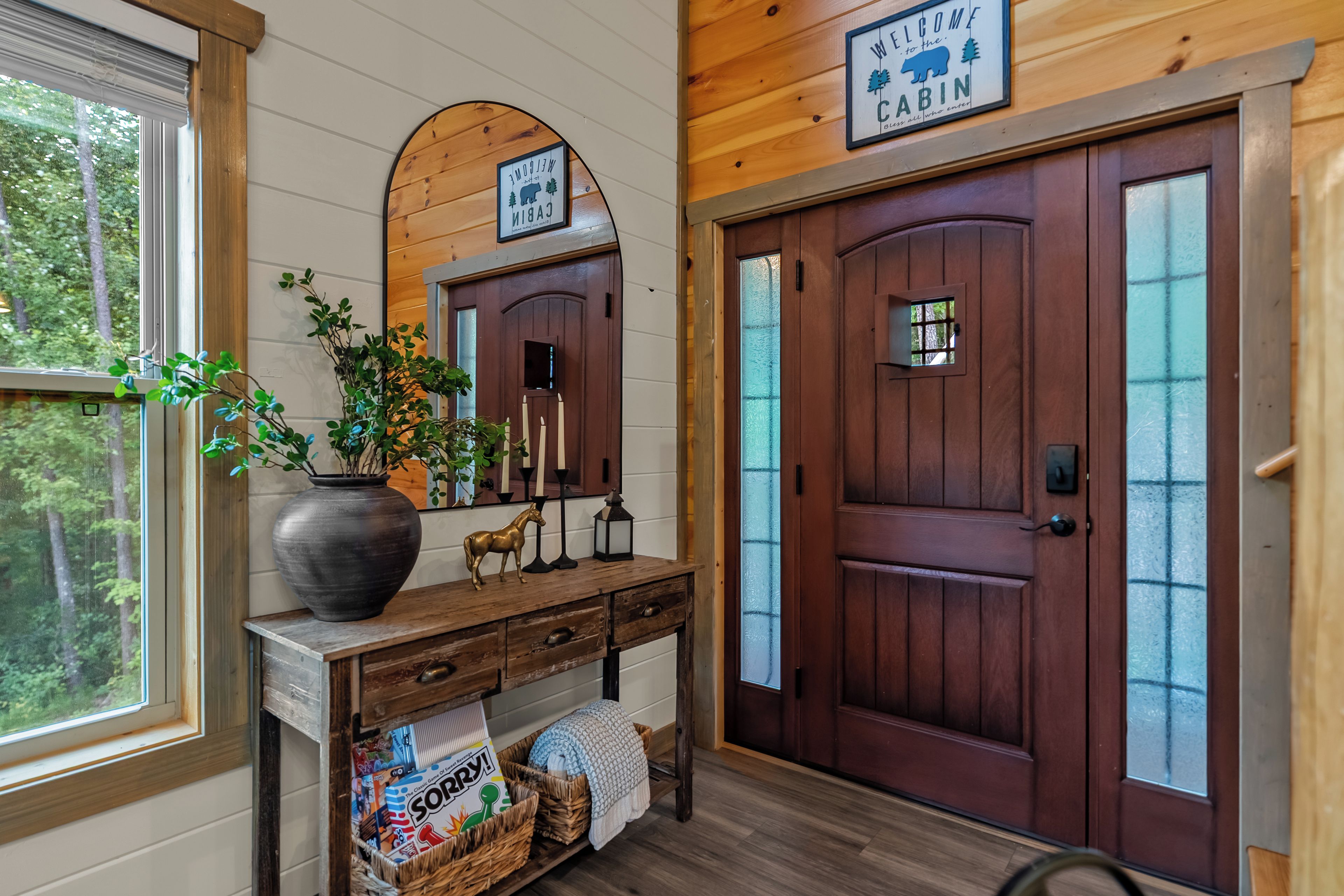 A cabin entrance with a wooden door, a welcome sign, a mirror, a vase with plants, and a basket of items.