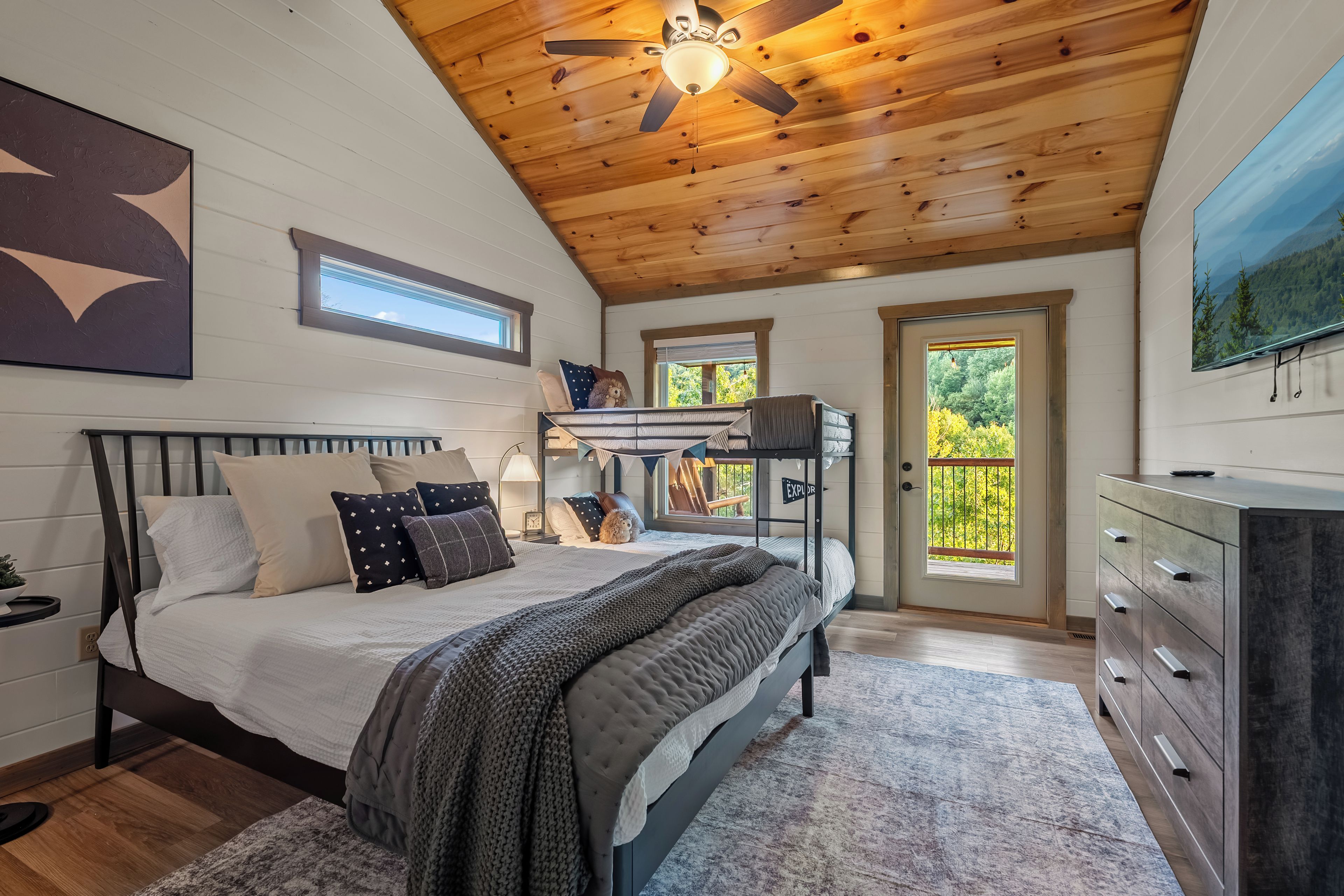 A bedroom with a bunk bed, a single bed, a ceiling fan, a large window, and a painting on the wall.