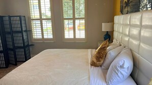 Een slaapkamer met een bed, kussens, een nachtkastje, een lamp en een raam met jaloezieën.