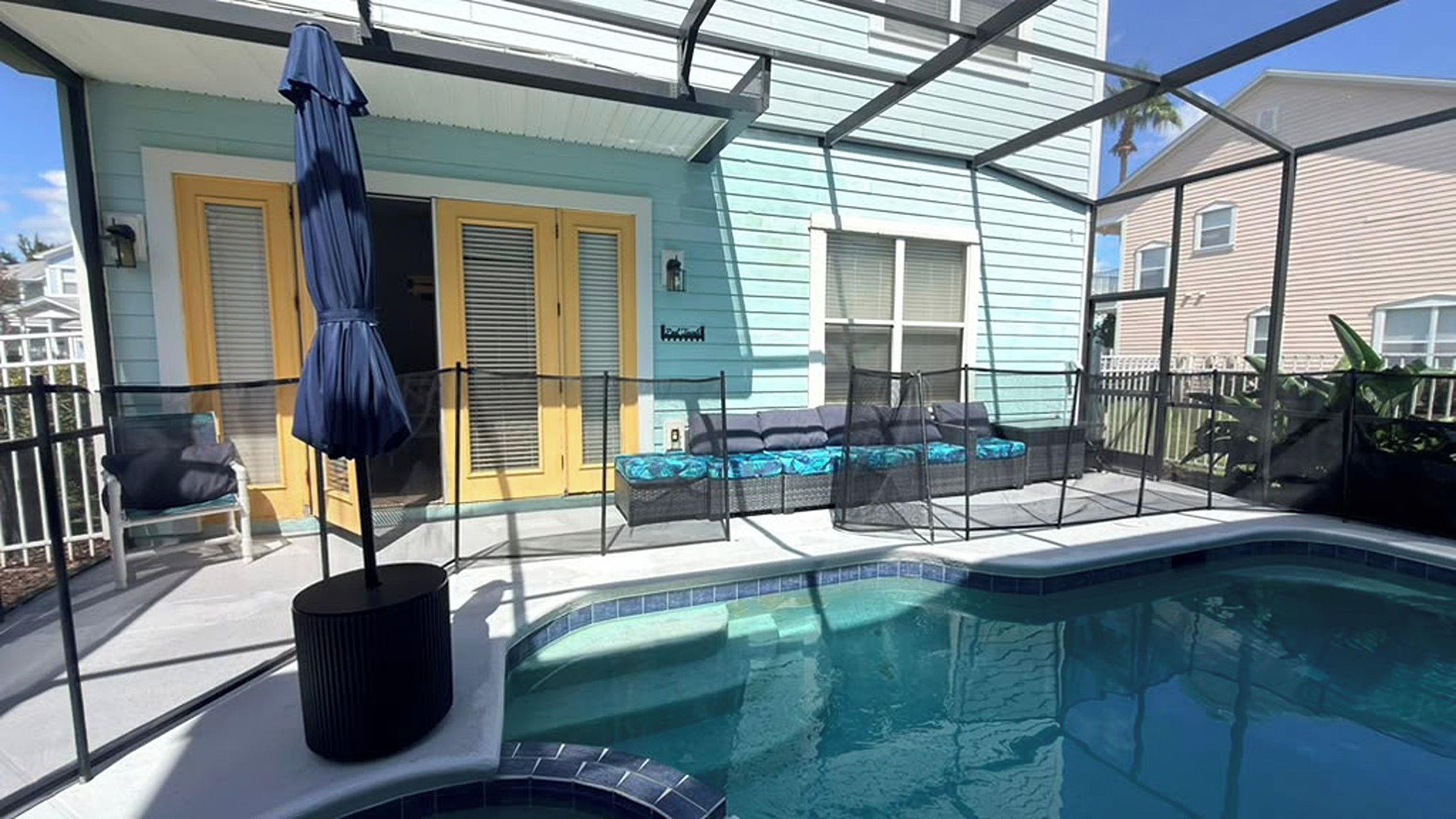 A swimming pool with a sunshade, a covered seating area with cushions, and a house with yellow shutters.