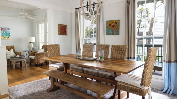 Un comedor con una mesa de madera, sillas de mimbre y un banco. Hay cortinas, una araña y obras de arte en las paredes.