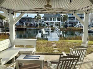 A covered outdoor area with white wooden furniture, a dock extending over water, and a view of houses in the background. - Cana Front Beauty close to the Heart of OIB! (Ocean Isle Beach)
