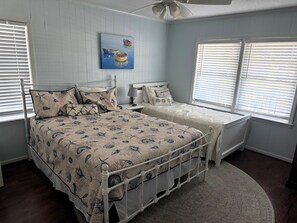 A bedroom with a bed, a nightstand, a lamp, a painting, and a window with blinds. - Cana Front Beauty close to the Heart of OIB! (Ocean Isle Beach)