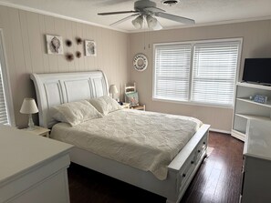 Een slaapkamer met een bed, een plafondventilator, een raam met zonwering, een televisie op een plank en een wandklok.