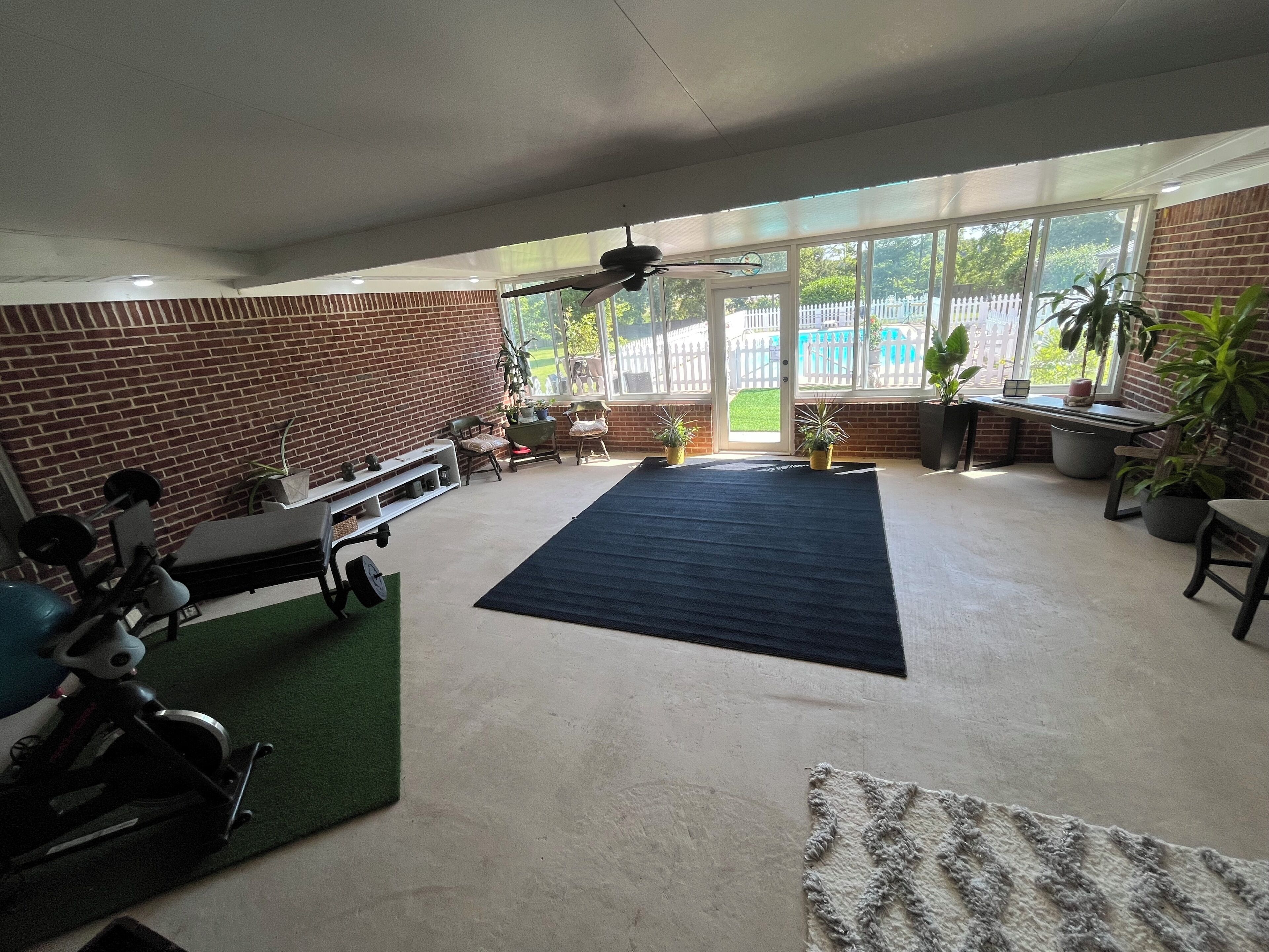 Ein geräumiges Zimmer mit großem Trainingsbereich, Deckenventilator und Blick auf einen eingezäunten Garten durch große Fenster.