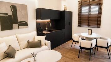 Un espacio habitable moderno con un sofá, una mesa de comedor redonda con sillas, una zona de cocina con un lavamanos y grifo, y una ventana con persianas.