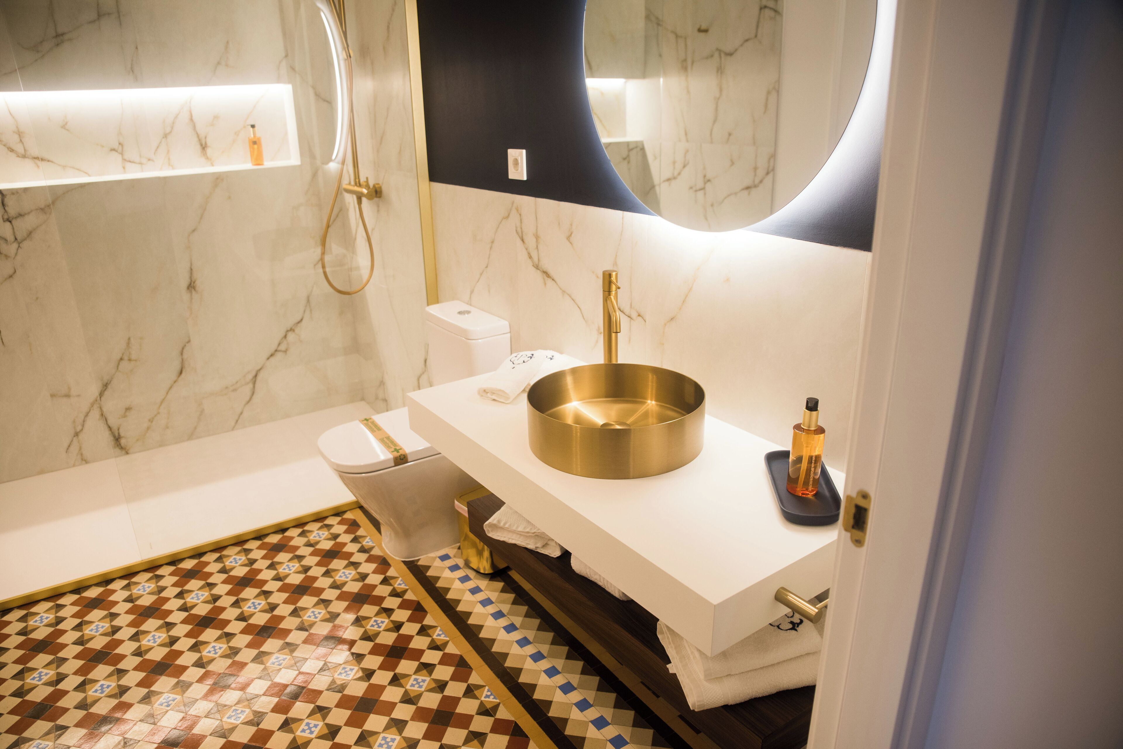 Un baño con pared de mármol, un lavamanos blanco, un grifo dorado y un suelo estampado.