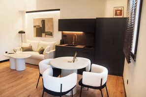 Un espacio habitable compacto con sofá, una mesa de centro, una mesa de comedor con sillas, una zona de cocina con fregadero y cafetera, y un cuadro en la pared.