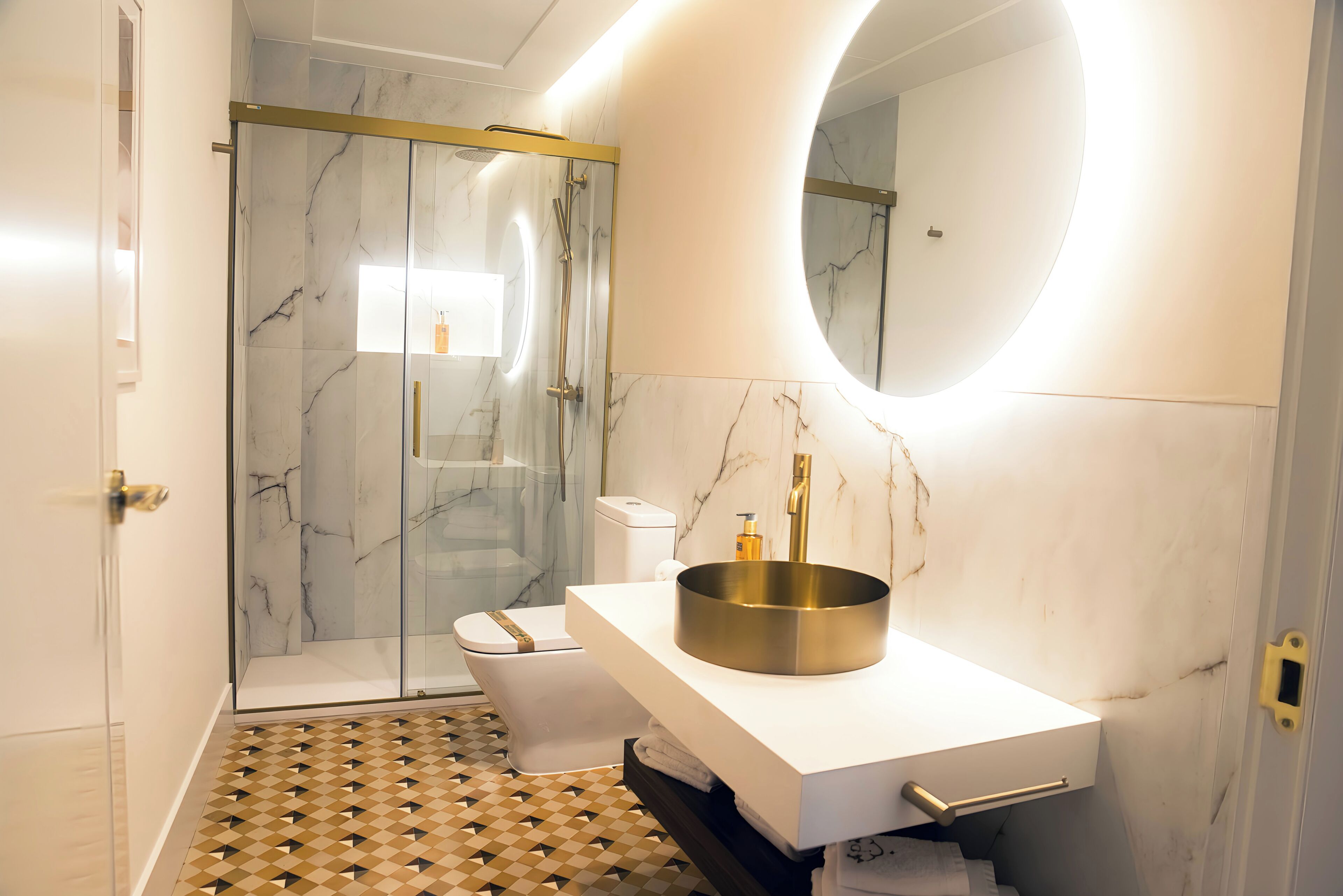 Un baño moderno con cabina de ducha de vidrio, lavabo blanco con lavamanos dorado y suelo de mármol.