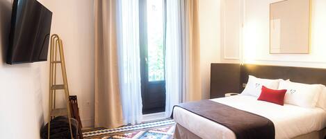 Quarto de hotel com cama, televisão fixada na parede, janela com cortinas e chão estampado.