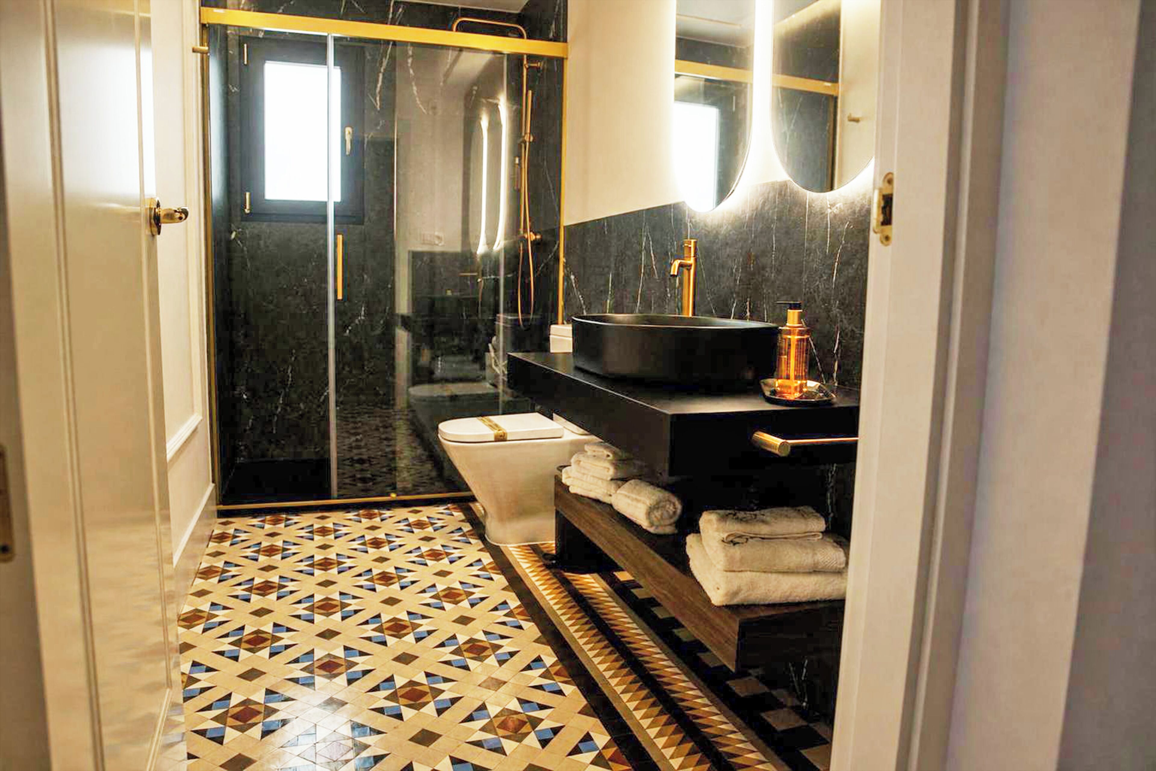 Un baño con ducha de vidrio, inodoro, lavamanos con espejo y suelo estampado.