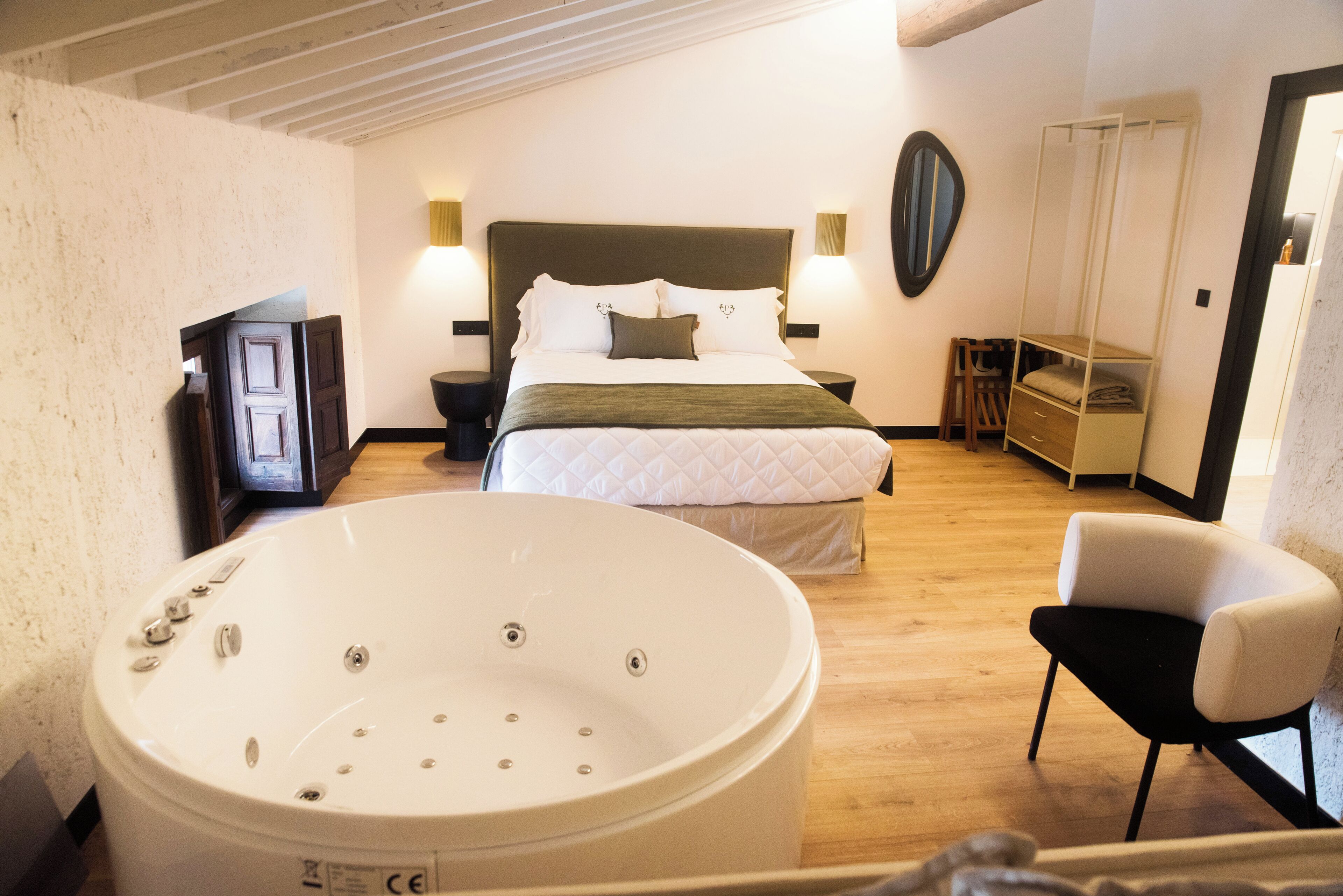 Habitación de hotel con una bañera grande, una cama con ropa blanca, suelo de madera, una silla y un armario.