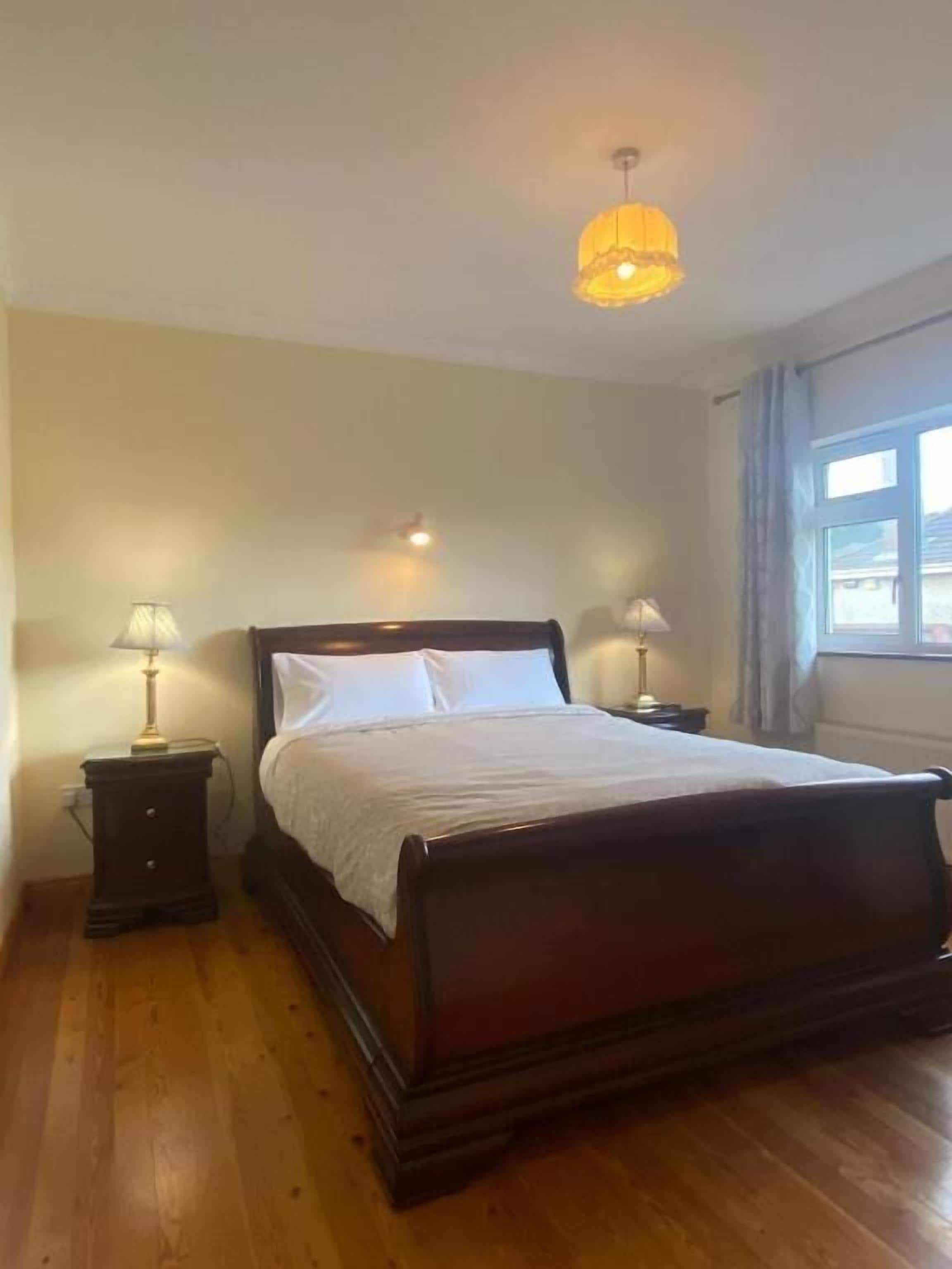 A bedroom with a wooden bed, two bedside tables with lamps, a ceiling light fixture, and a window with curtains.