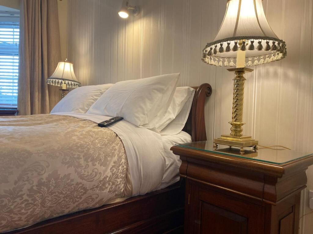 A neatly made bed with a remote control, a glass side table with a lamp, and a wooden nightstand.