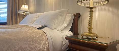 A neatly made bed with a remote control, a glass side table with a lamp, and a wooden nightstand.